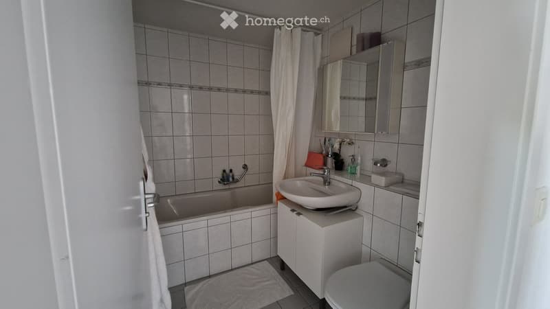 Bathroom with a bathtub, sink, toilet, mirror, and cabinet. The walls are tiled, and there is a shower curtain in the background.