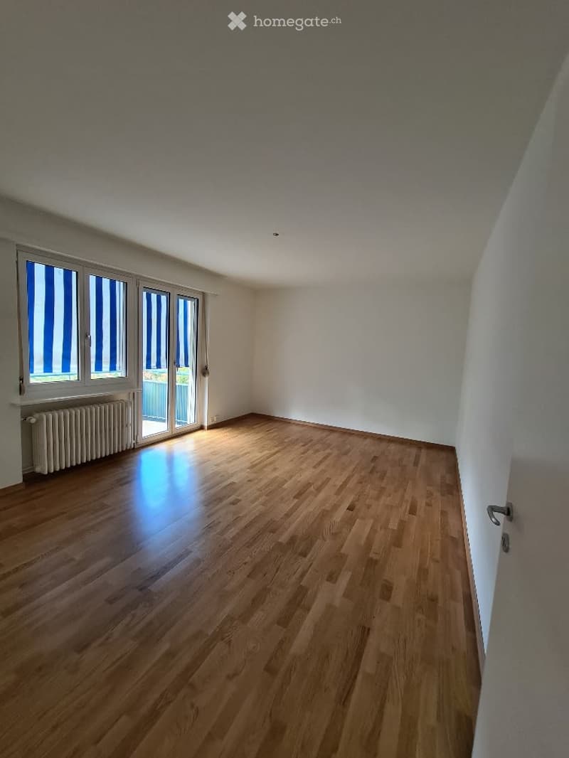 Empty room with wooden flooring, white walls, large windows with blinds, and a radiator under one window.