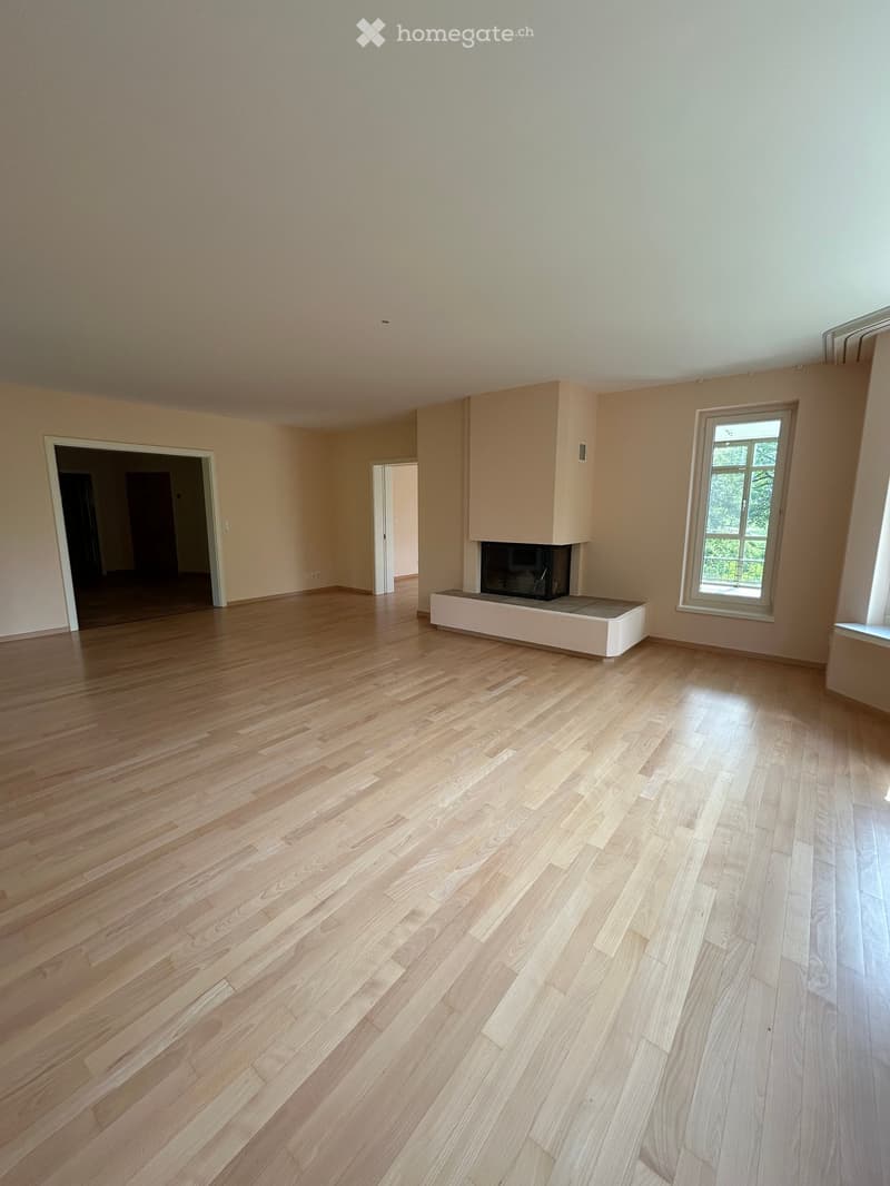 Empty room with light wooden flooring, white walls, and a built-in fireplace with a white surround. There are two doors and a window with a view of greenery.