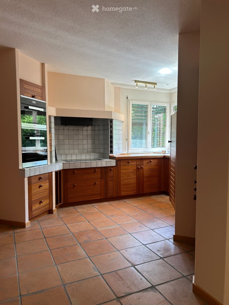 Bright kitchen with wooden cabinets, tiled floor, and a large window overlooking the garden. Features an oven, countertop, and sink.