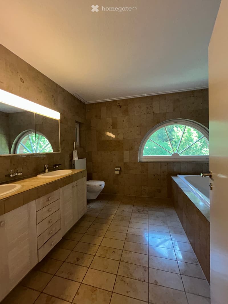 Bathroom with beige tiled walls and floor, double sink vanity, bathtub, toilet, and large arched windows allowing natural light.