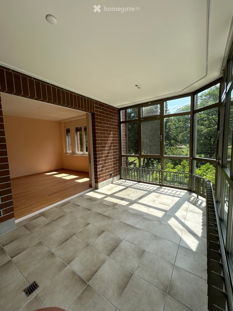 Empty room with tiled floor, wooden flooring in adjacent area, large windows with metal railings, and a view of greenery outside.