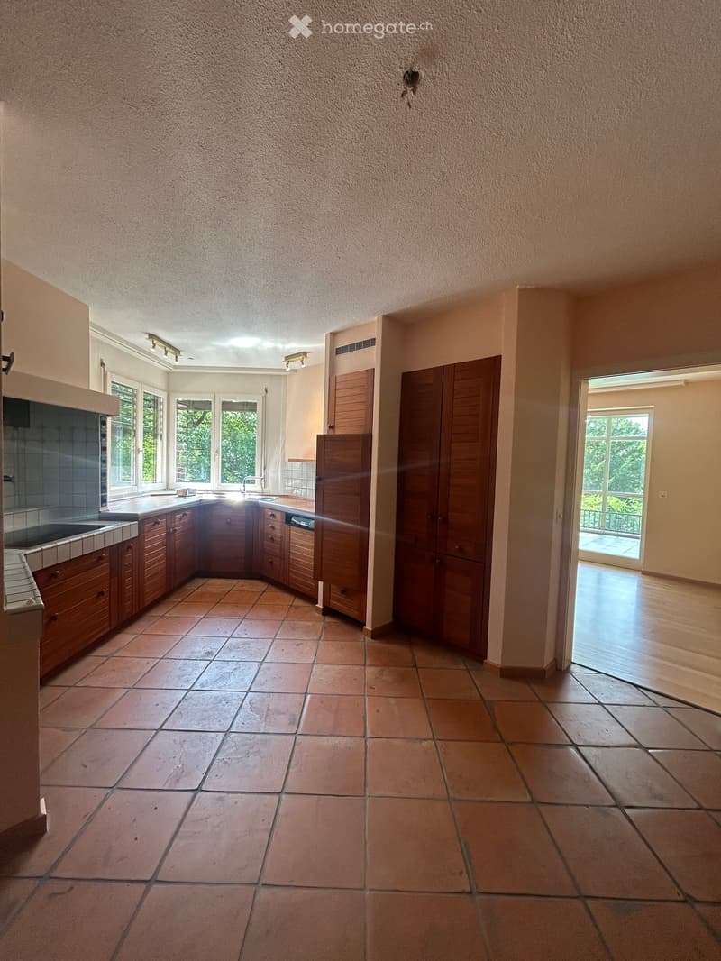 Empty kitchen with wooden cabinets, tiled floor, and large windows. Features a sink, faucet, and a dishwasher.