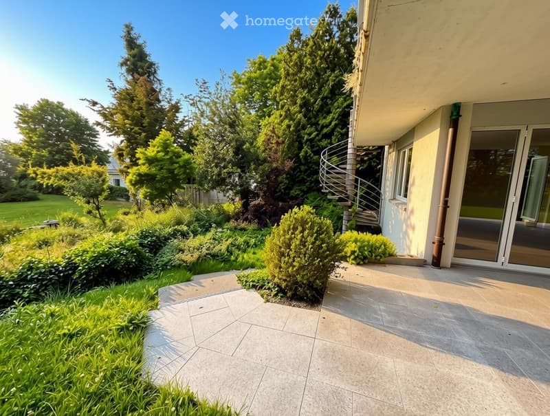 Well-maintained garden with a variety of trees, shrubs, and a paved patio area adjacent to a house with a spiral staircase leading to an upper level.