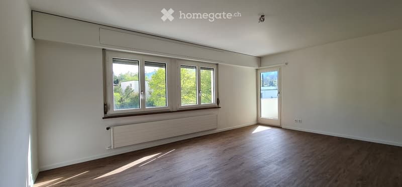 Empty room with wooden flooring, large windows, and a sliding glass door leading to a balcony.
