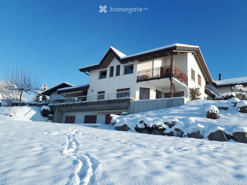 A two-story white house with snow-covered roof and balconies, surrounded by snow-covered ground and trees.