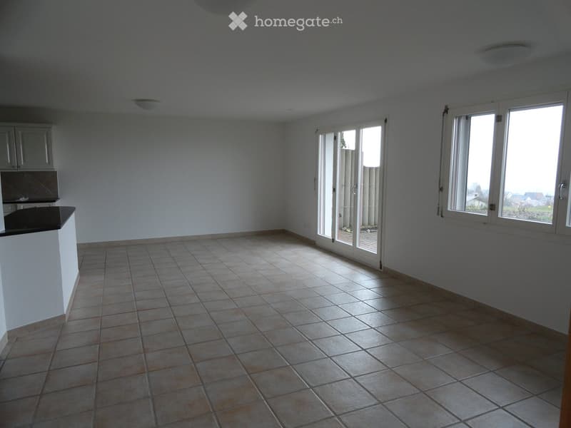 Empty kitchen with tiled floor, white walls, and large windows. Minimal cabinetry visible on one side.