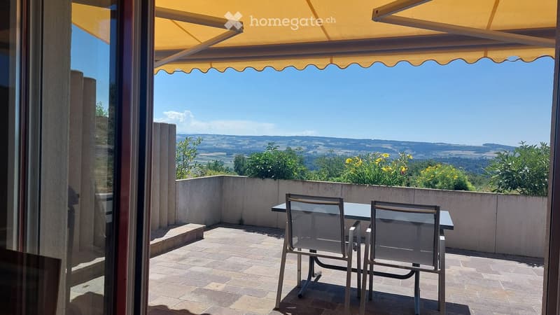 Balcony with a table and two chairs, overlooking a scenic countryside view with a yellow awning providing shade.