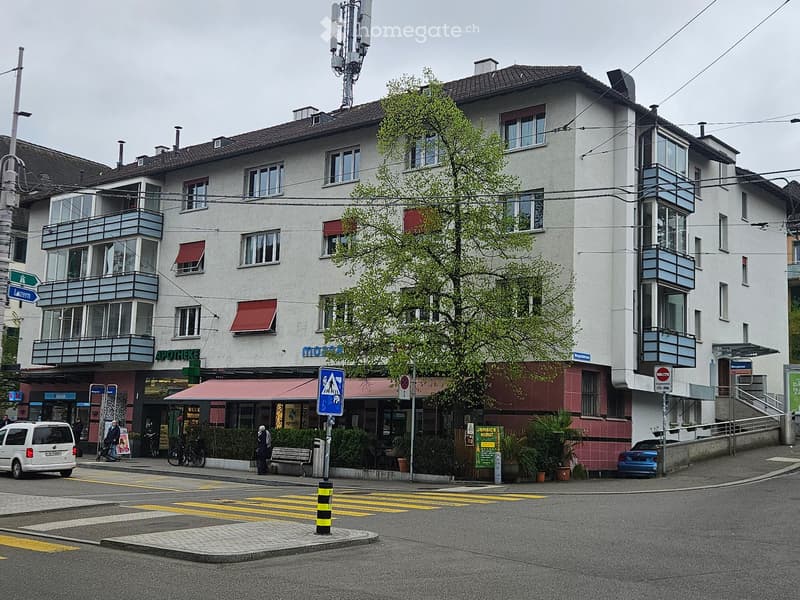 Multi-story building with balconies, storefronts, and signage for a pharmacy and other businesses. The ground floor includes a restaurant with outdoor seating, and there are parked cars and pedestrians visible.