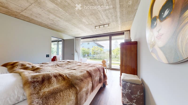 Modern bedroom with a large bed covered in a fur throw, a nightstand, a chair, a stool, a wooden cabinet, and a large painting of a woman with sunglasses on the wall. The room features large windows with curtains and a view of the garden.