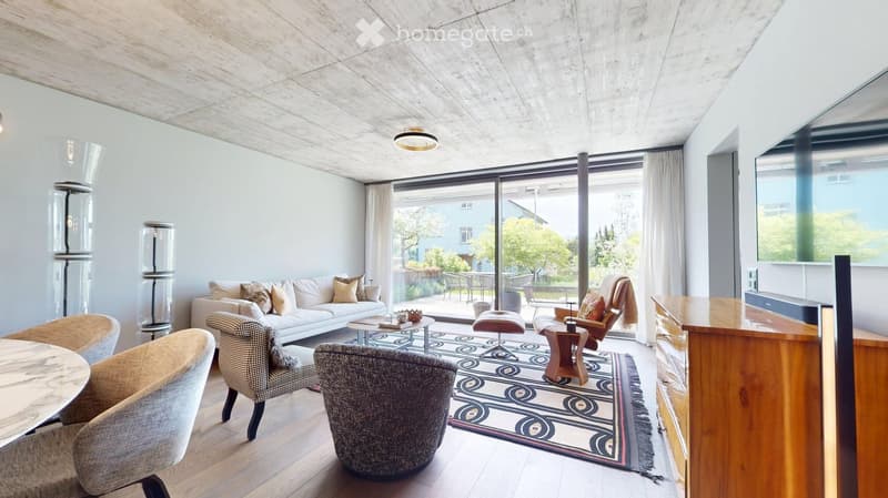 Modern living room with white walls, wooden floor, large windows, and a view of the garden. Features a white sofa, coffee table, several chairs, a stool, and a patterned rug.