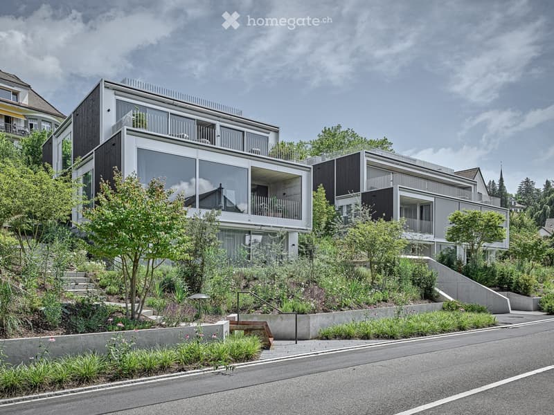 Modern multi-story building with large glass windows and balconies, situated on a raised ground floor with landscaped surroundings.