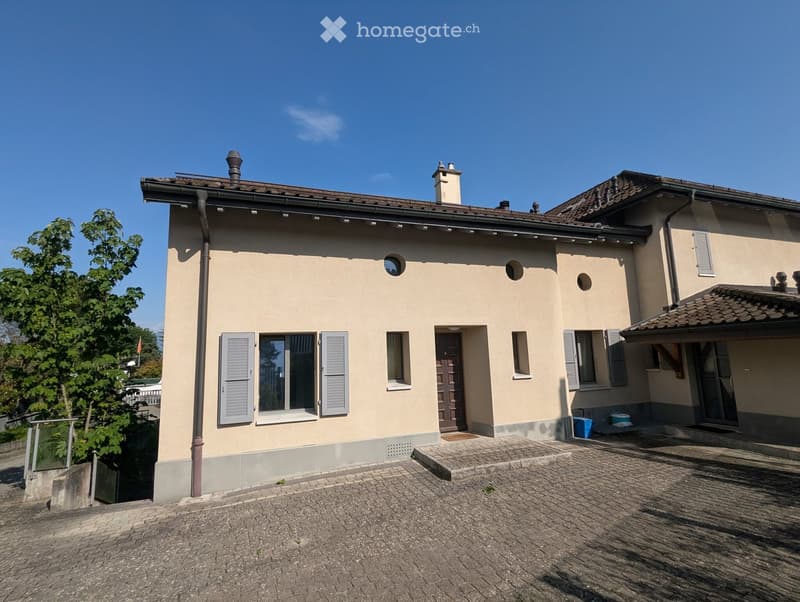Two-story house with beige exterior, multiple windows with shutters, a central entrance door, and a tiled driveway.