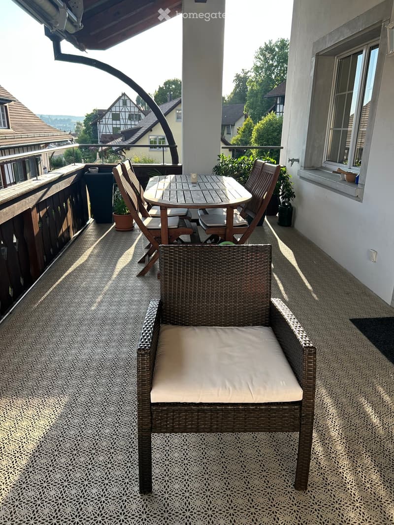 Balcony with a wicker chair, a wooden table, and two folding chairs, overlooking a residential area with traditional houses.