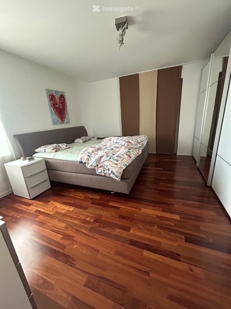 Modern bedroom with wooden flooring, a large bed with a headboard, nightstand, and a painting on the wall.