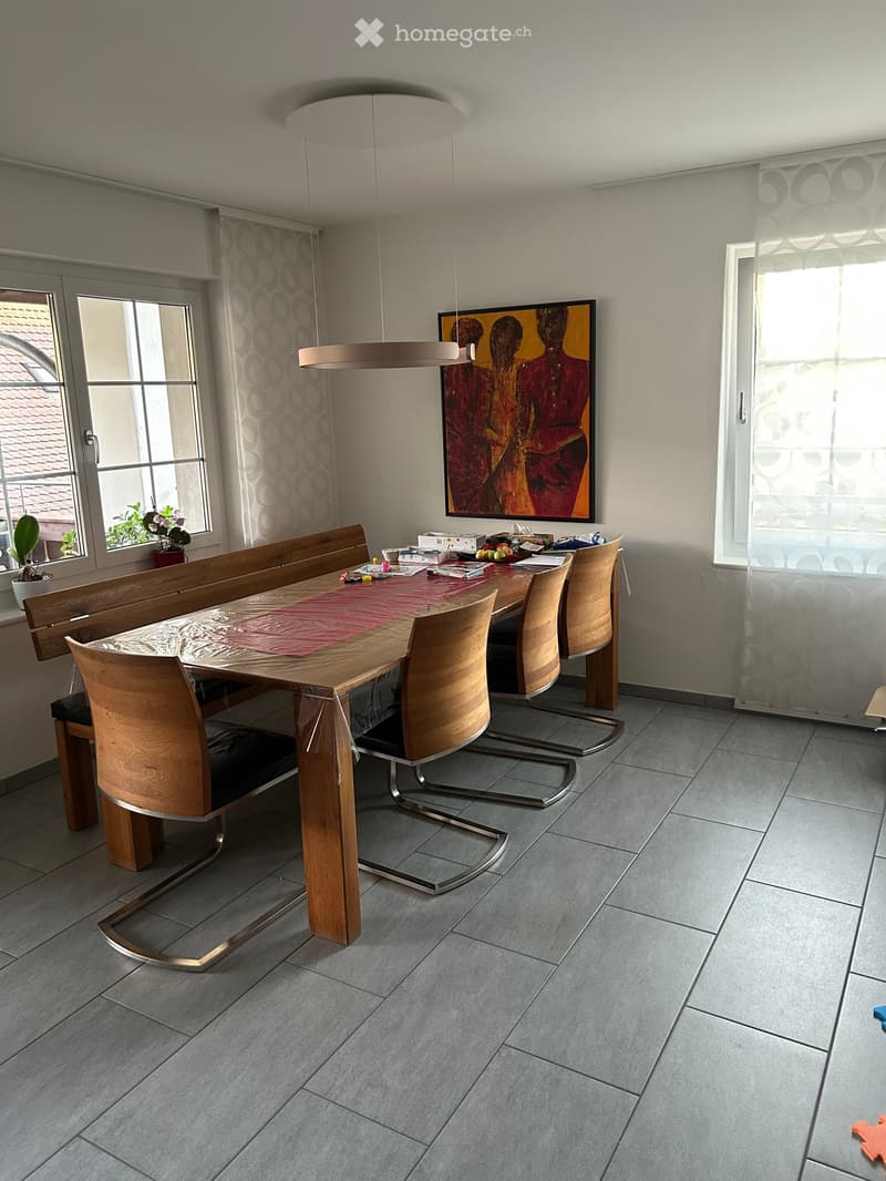 Dining room with a wooden dining table, six chairs, pendant light, and a painting on the wall. The room has large windows with curtains and a potted plant on the windowsill.