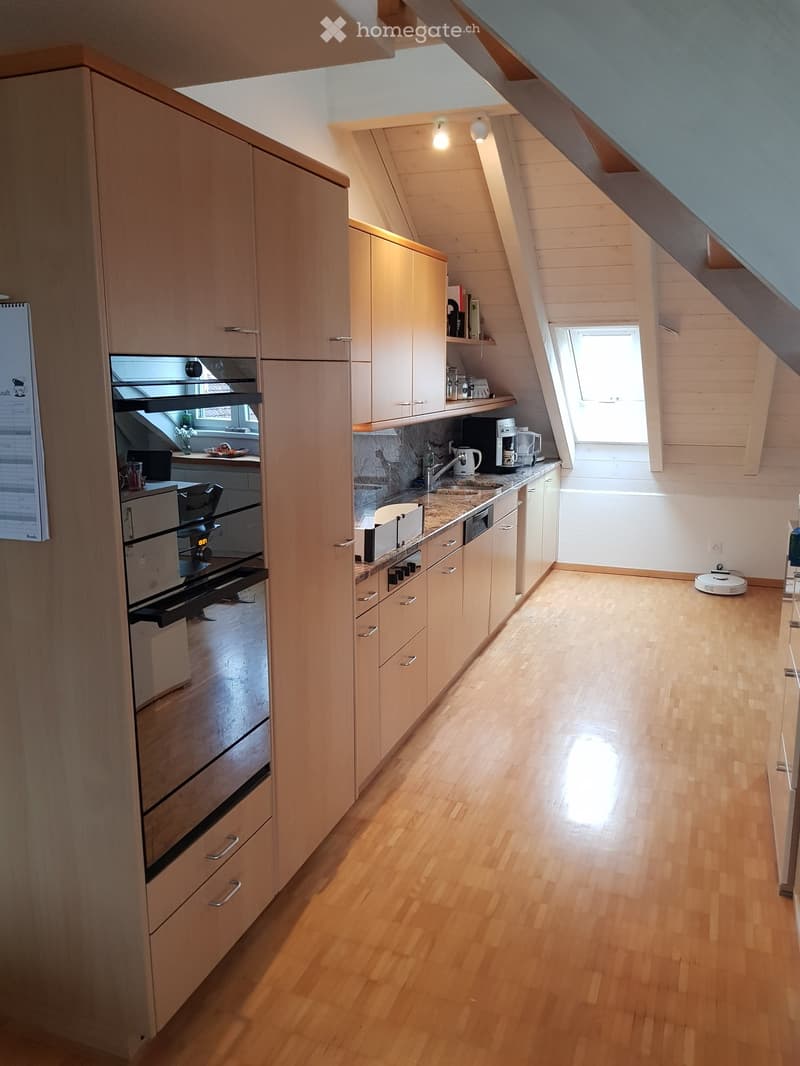 Modern kitchen with light wooden cabinets, stainless steel appliances, and a marble countertop. The space features a built-in oven, a sink with a faucet, and a coffee maker on the counter. There is a small table with a laptop and a chair in the corner, an