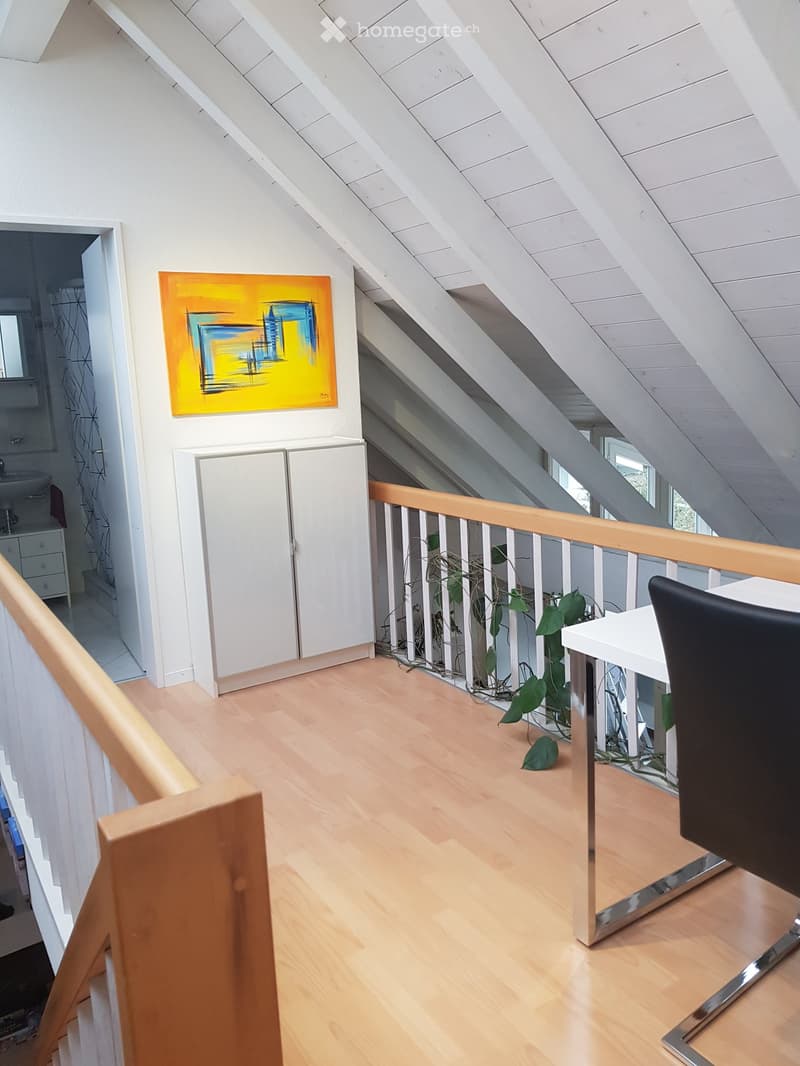 Attic room with wooden floors, slanted white ceiling, white cabinets, a black chair, a white table, a potted plant, and a colorful abstract painting on the wall.