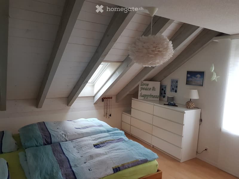 Cozy attic bedroom with slanted wooden ceilings, skylight windows, a double bed with blue bedding, a white dresser, and decorative items including a hanging white pom-pom light fixture.
