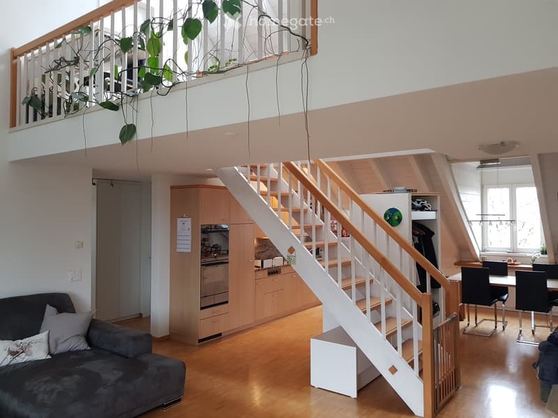 Open-plan living and dining area with a staircase leading to an upper level, featuring a sofa, dining table with chairs, kitchen cabinets, and a potted plant on the upper level.