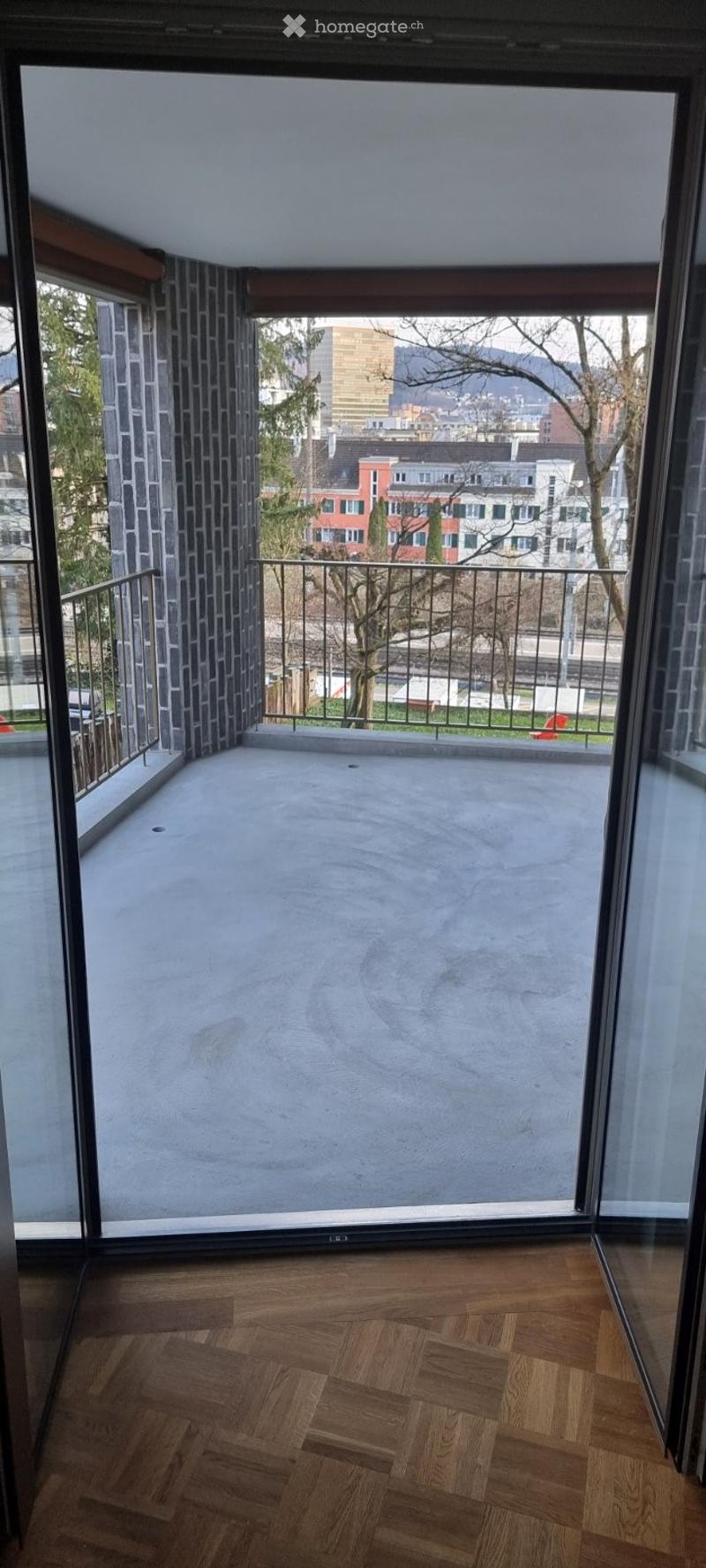 Spacious balcony with wooden flooring, large glass doors, metal railing, and a view of the city and trees.