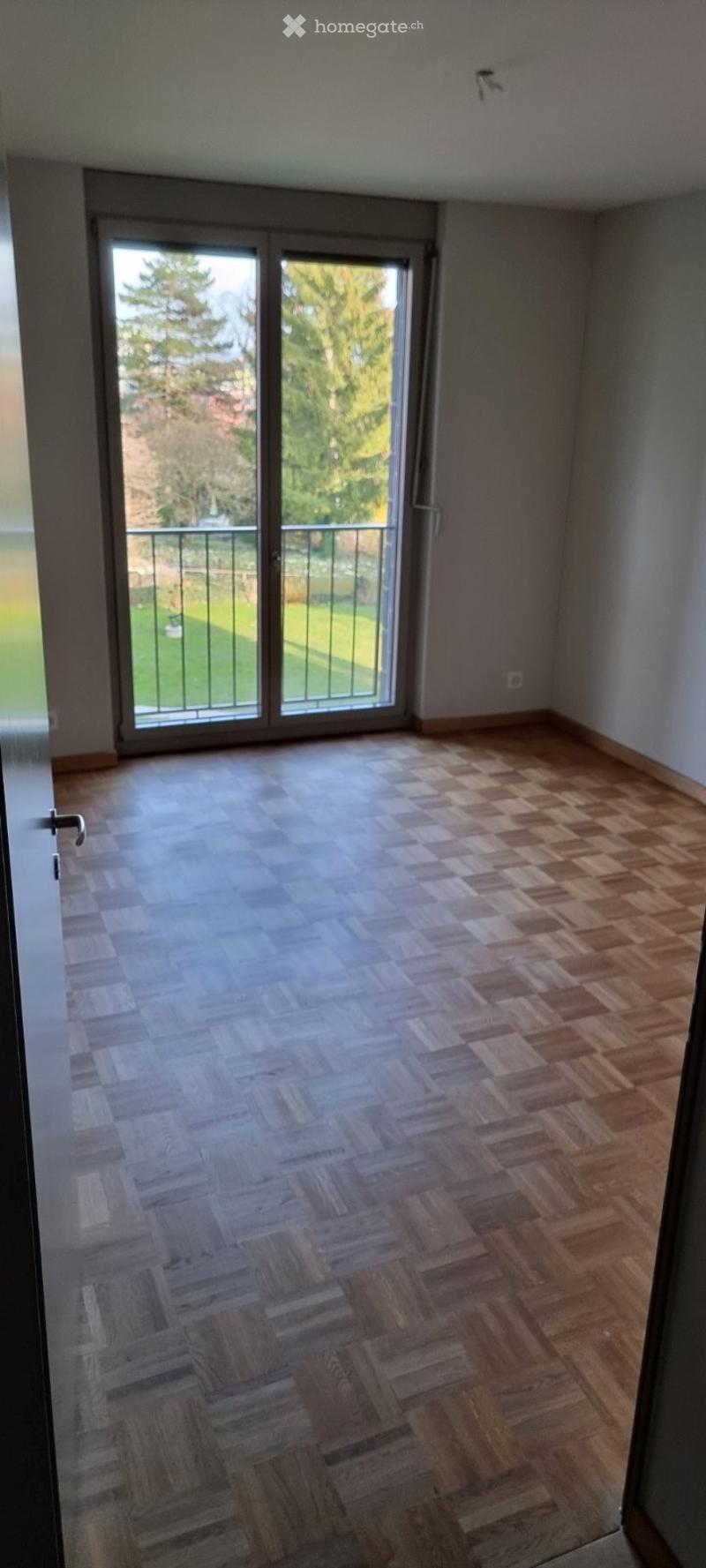 Empty room with wooden herringbone flooring, large sliding glass door leading to a balcony with a view of greenery.