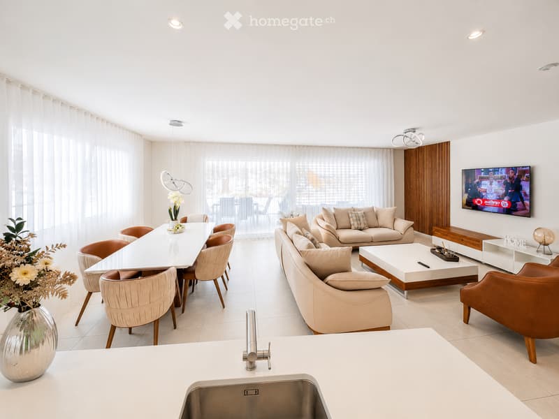 Modern open-plan living and dining area with a white dining table, beige chairs, a large sectional sofa, a coffee table, and a television. The space features large windows with sheer curtains and a sleek kitchen counter with a sink.