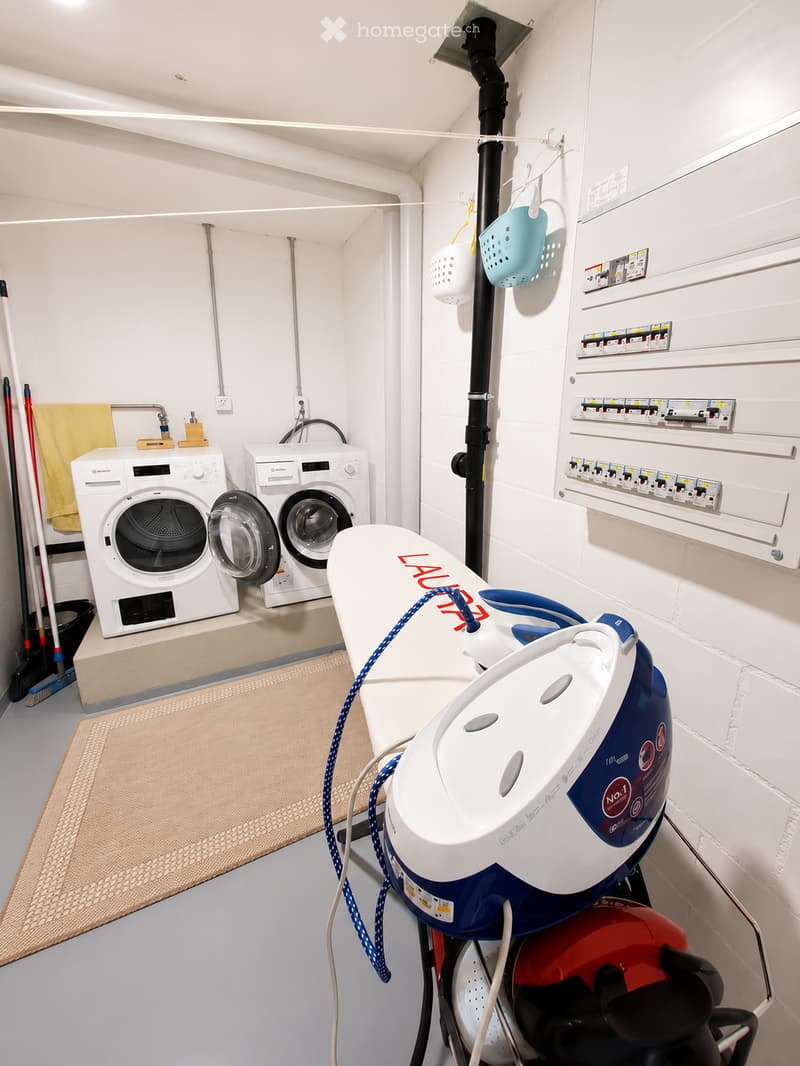 A compact utility room with a washing machine, dishwasher, ironing board, and cleaning supplies.