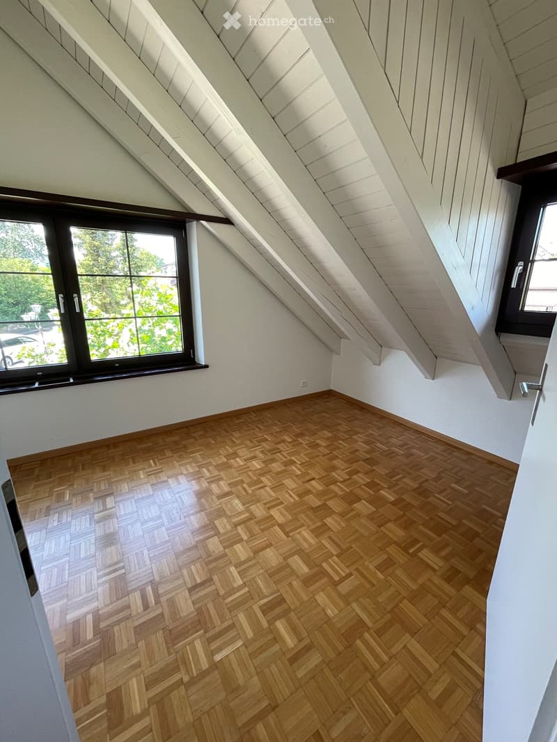Empty attic room with wooden herringbone flooring, white walls, and exposed wooden beams on the ceiling. Two windows provide natural light, one with a view of greenery outside.