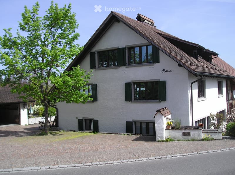 Two-story house with white walls, green shutters, and a brown tiled roof. Features include a garage, a driveway, and a small garden area with plants and a bicycle.