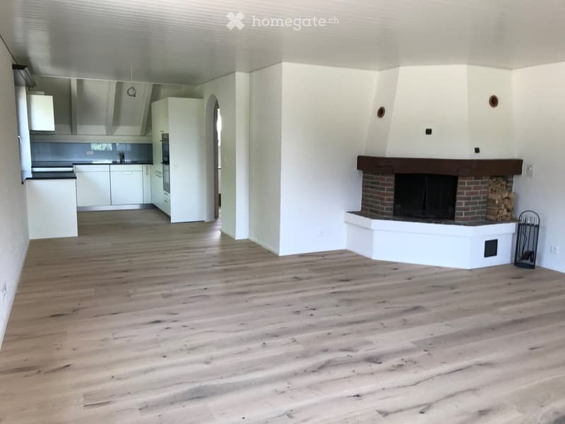 Open-plan living space with light wooden flooring, white walls, and a brick fireplace with a wood stack nearby. The kitchen area features white cabinets and stainless steel appliances.