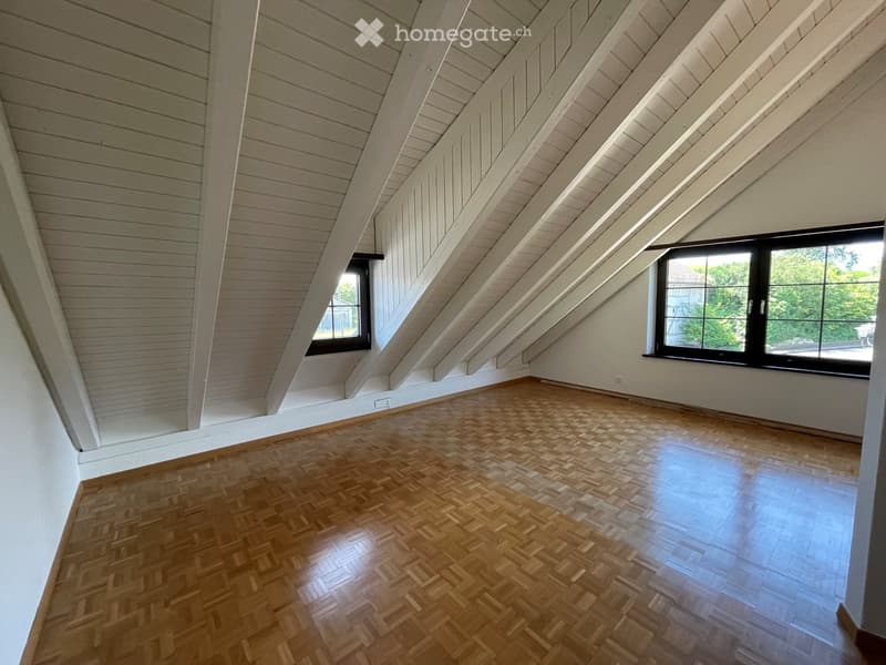 Empty attic room with wooden herringbone flooring, slanted wooden ceiling, and two windows offering natural light.