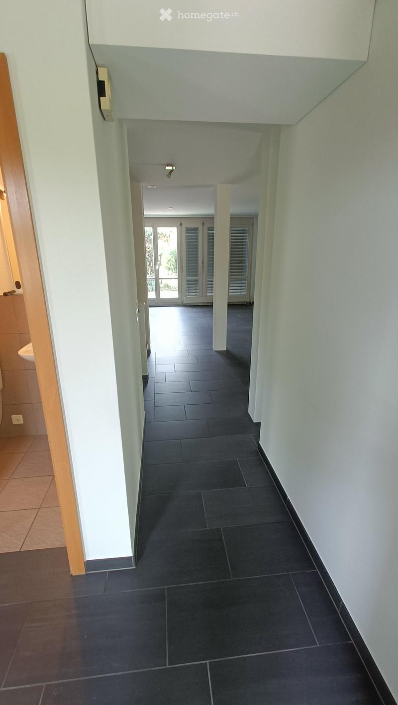A long, narrow hallway with dark tiled flooring, light-colored walls, and a wooden door frame on the left side. The hallway leads to a bright area with large glass doors and shutters at the end.