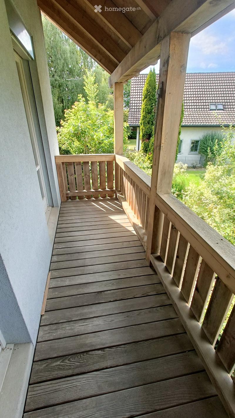 Wooden balcony with a railing, overlooking a garden area with trees and a neighboring house.
