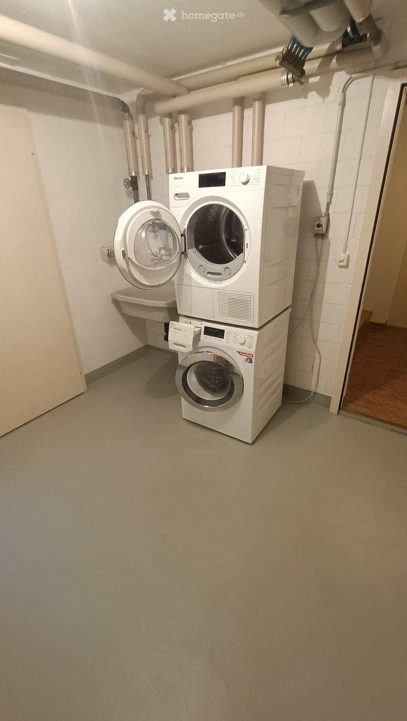 Laundry room with stacked white washing machine and dryer, beige walls, grey floor, and exposed pipes.