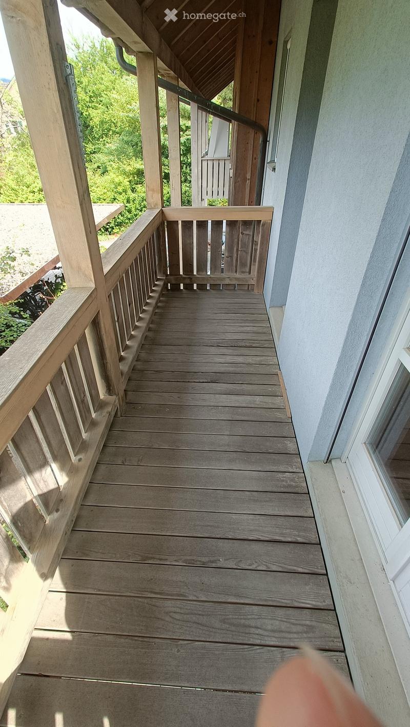 Wooden balcony with railings, leading to a green outdoor area.