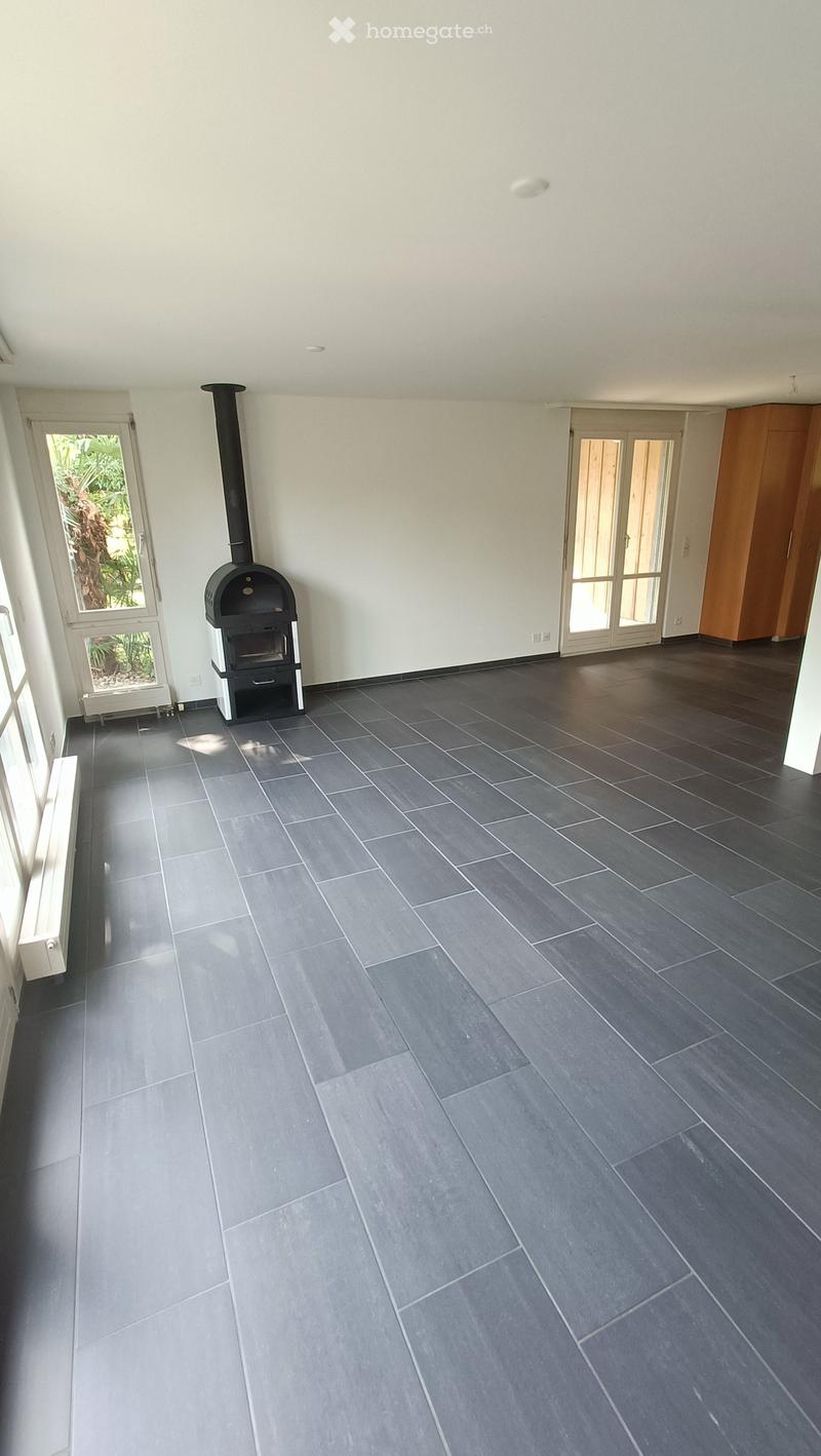 Empty room with dark grey tiled floor, white walls, large windows, and a black wood-burning stove with chimney.