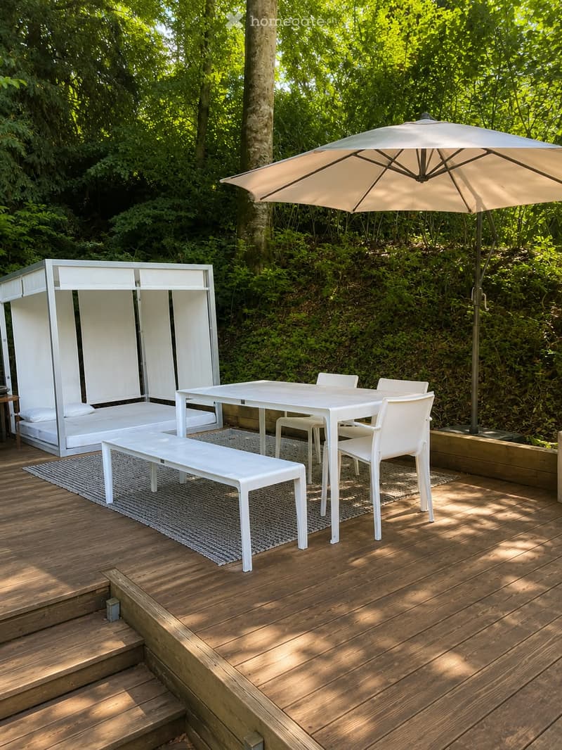 A wooden deck area with a white table, four white chairs, a white bench, a large umbrella, and a small white bed with pillows.