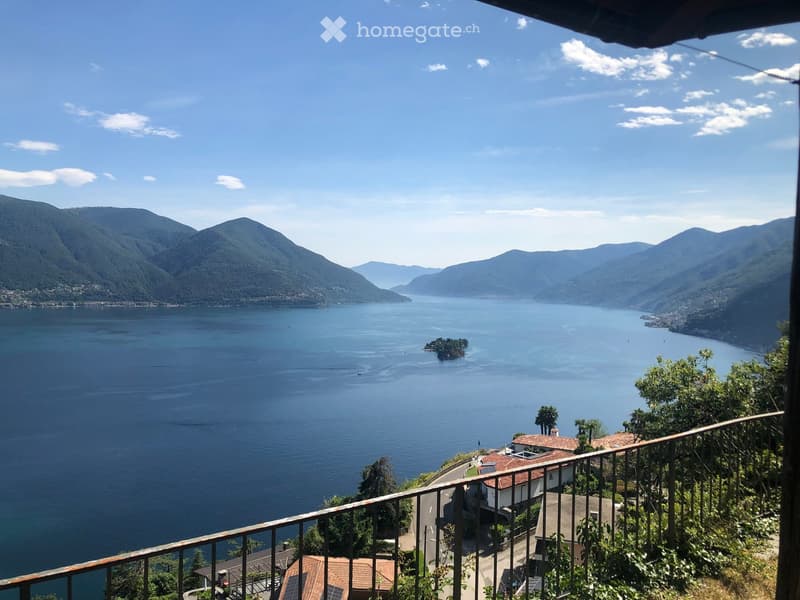 Balcony overlooking a serene lake surrounded by mountains, with a clear blue sky and distant shoreline visible.