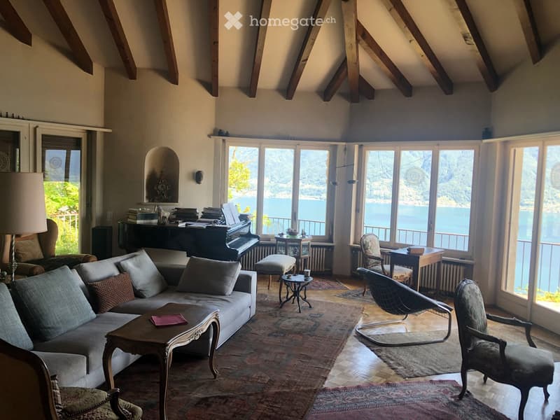 Spacious living room with large windows overlooking a scenic lake view, featuring a grey sofa, coffee table, chairs, piano, and decorative items.