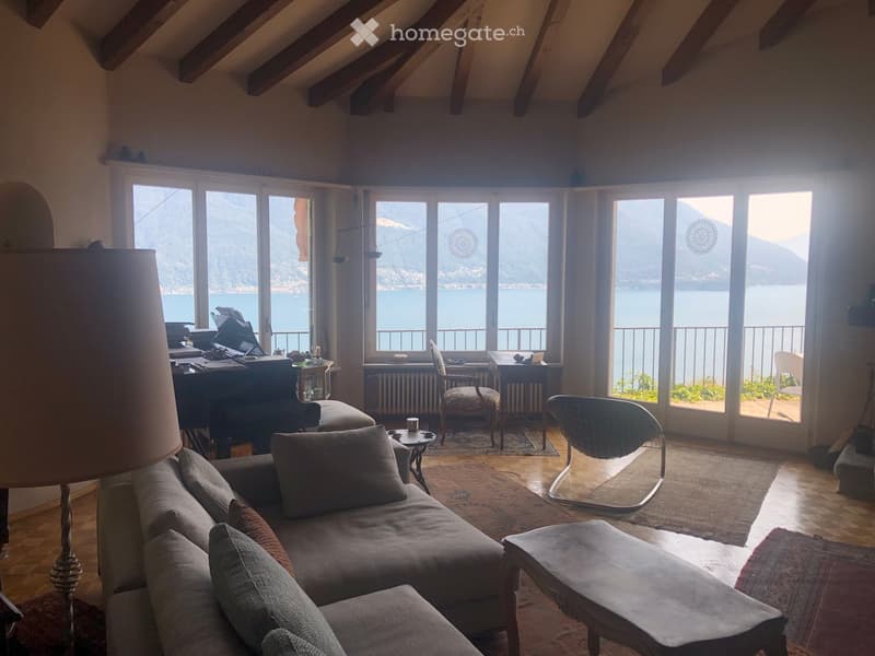 Bright living room with large windows overlooking a lake, featuring a sofa, coffee table, chairs, a desk, and a lamp.
