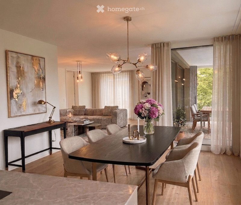 Modern living and dining area with a grey sofa, coffee table, dining table with chairs, table lamp, flower vase, and artwork on the wall.