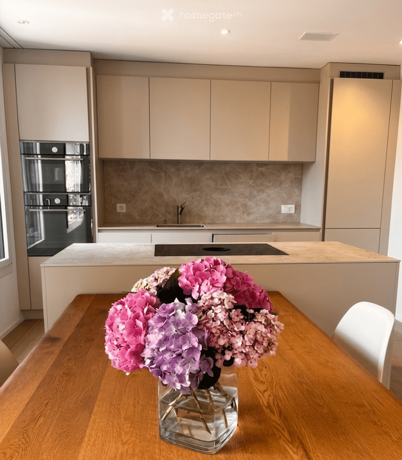 Modern kitchen with light-colored cabinets, a central island, and a vase of pink and purple flowers on the dining table.