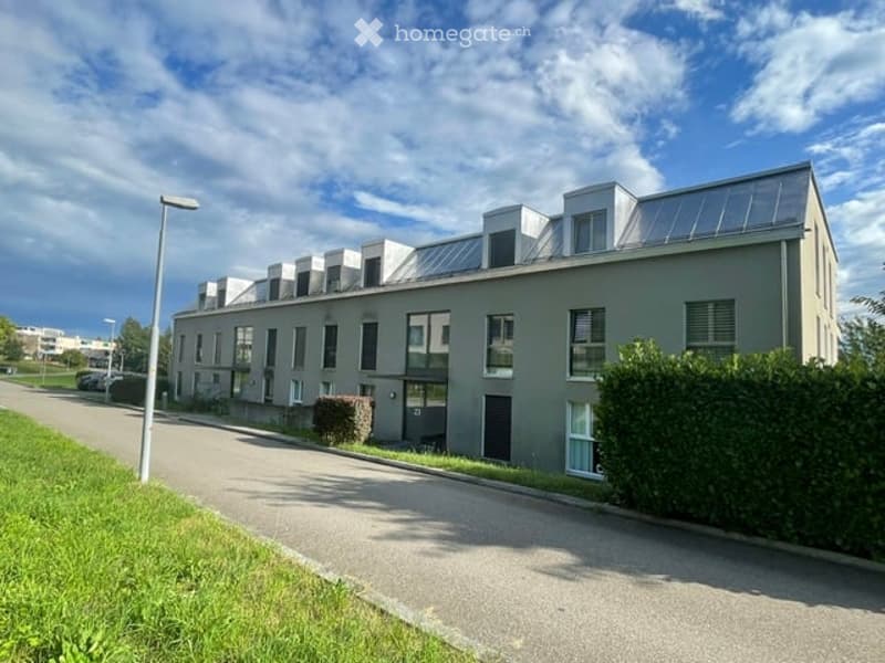 Modern multi-story building with flat roof and large windows, surrounded by greenery and a parking area.