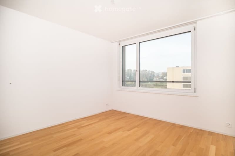 Empty room with white walls, wooden floor, and large window offering a view of the outside.
