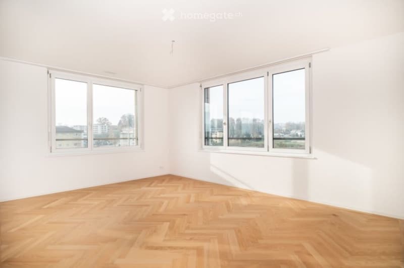 Empty room with white walls, wooden herringbone flooring, and large windows offering a view of the city and trees.