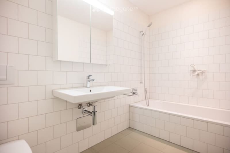 Modern bathroom with white tiled walls and floor, featuring a sink, bathtub, and mirror.