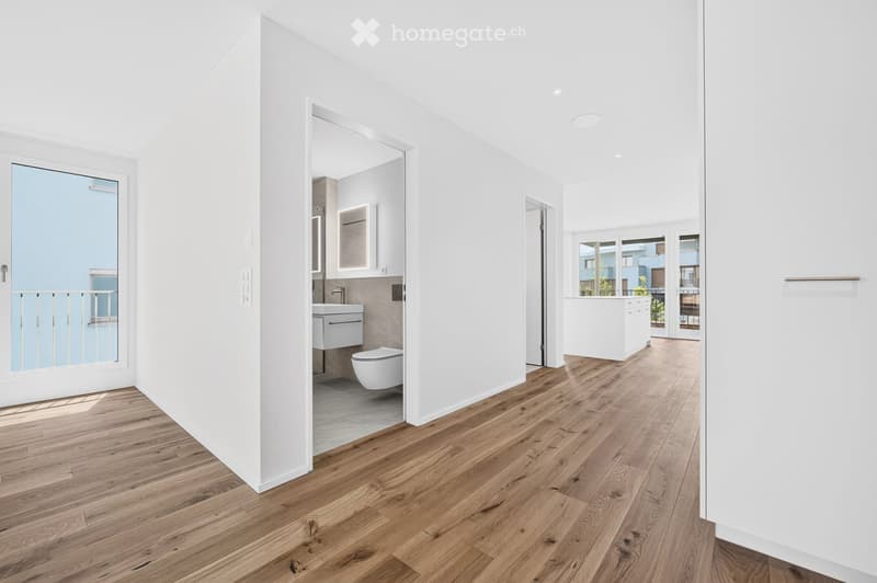 Bright, minimalist hallway with wooden flooring, white walls, and recessed lighting. Features a modern bathroom with a floating sink, toilet, and built-in cabinetry, accessible through an open doorway.