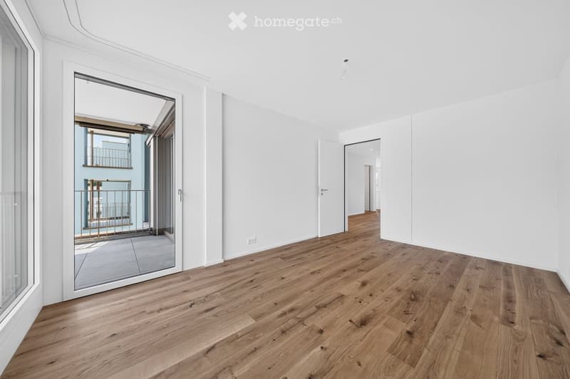 Empty room with light wooden flooring, white walls, and large windows. A sliding glass door leads to a balcony with a metal railing.