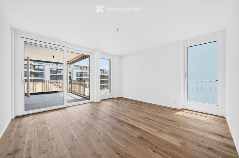 Empty room with light wooden flooring, white walls, large sliding glass doors leading to a balcony, and a view of a modern building complex.